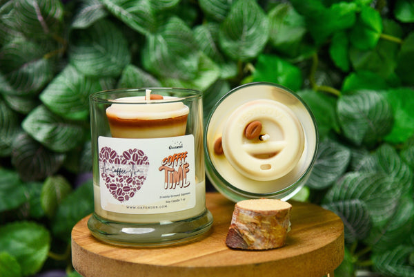 A glass jar candle labeled 'Coffee Time' with a cream-colored wax and a brown paper label, displayed next to its lid on a round wooden stand in front of a green leafy background.